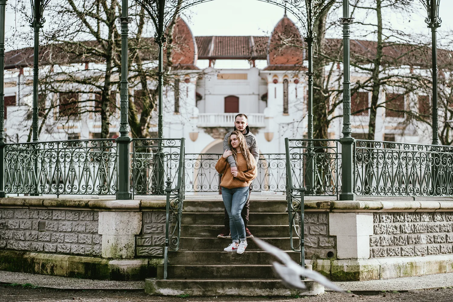 Un couple se tient étroitement sur les marches d'un kiosque en fer forgé dans un parc, avec un bâtiment blanc à l'arrière-plan, illustrant des moments intimes et authentiques de leurs vacances à Porto, typiques des photoreportages de mariage ou séances photo de famille dans les Landes; un oiseau en vol près du bas de l'image ajoute une touche dynamique et poétique à cette scène sereine.