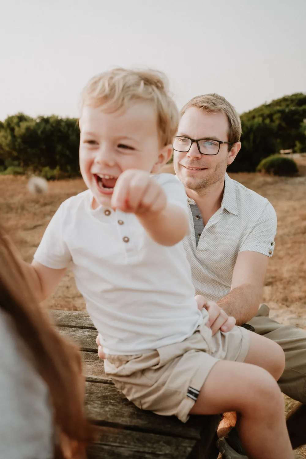 Un jeune garçon souriant vêtu d'une chemise blanche pointe vers l'avant tandis qu'un adulte portant des lunettes et une chemise de couleur claire est assis derrière lui sur un banc en bois, illustrant parfaitement la chaleur des moments familiaux capturés par un photographe. de famille dans les Landes.