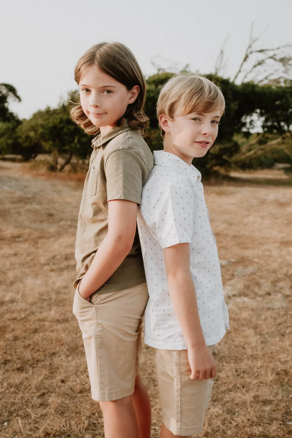 Dans cette touchante photographie de famille capturée par un talentueux photographe de mariage et famille dans les Landes, deux jeunes garçons se tiennent dos à dos dans un champ aride avec une rangée d'arbres en arrière-plan, vêtus tous deux de shorts et de chemises. à manches courtes, illustrant parfaitement l'essence des souvenirs d'été en plein air.