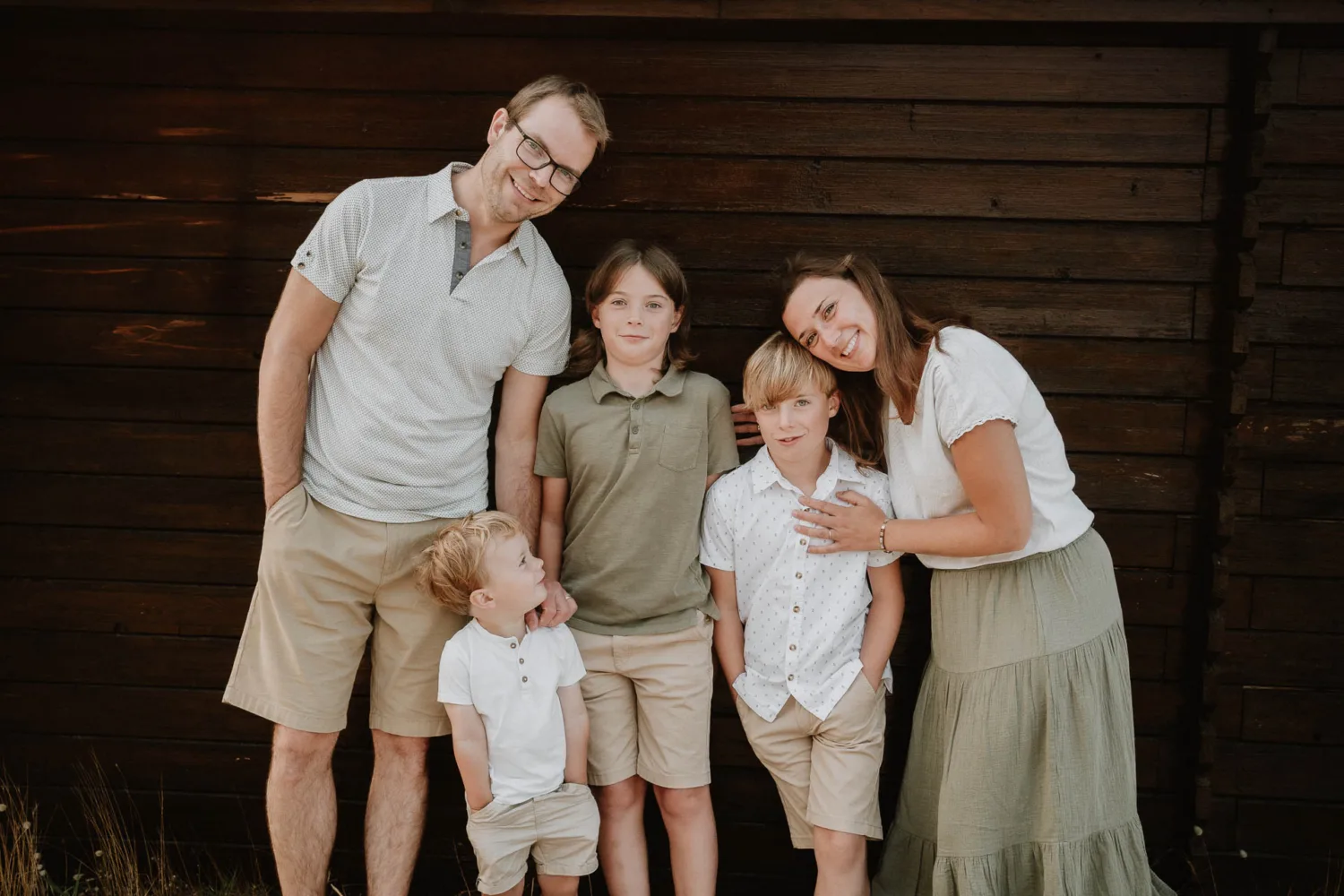 Une famille de cinq personnes se tient devant un mur en bois sombre, magnifiquement capturée par un photographe de famille dans les Landes ; les parents sont positionnés de chaque côté de leurs trois jeunes garçons, tous vêtus de vêtements d'été décontractés.