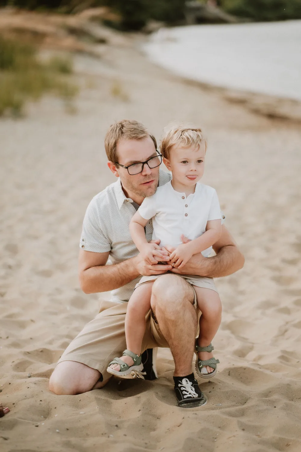 Sur cette magnifique photo de famille capturée par un photographe professionnel spécialisé dans les mariages et les familles dans les Landes, un homme adulte se tient à genoux sur le sable de la plage, tenant tendrement un jeune enfant sur ses genoux, tous deux regardant légèrement vers la gauche, offrant une composition parfaite qui témoigne de l'intimité et du bonheur familial typique d'une séance photo sur le littoral landais.