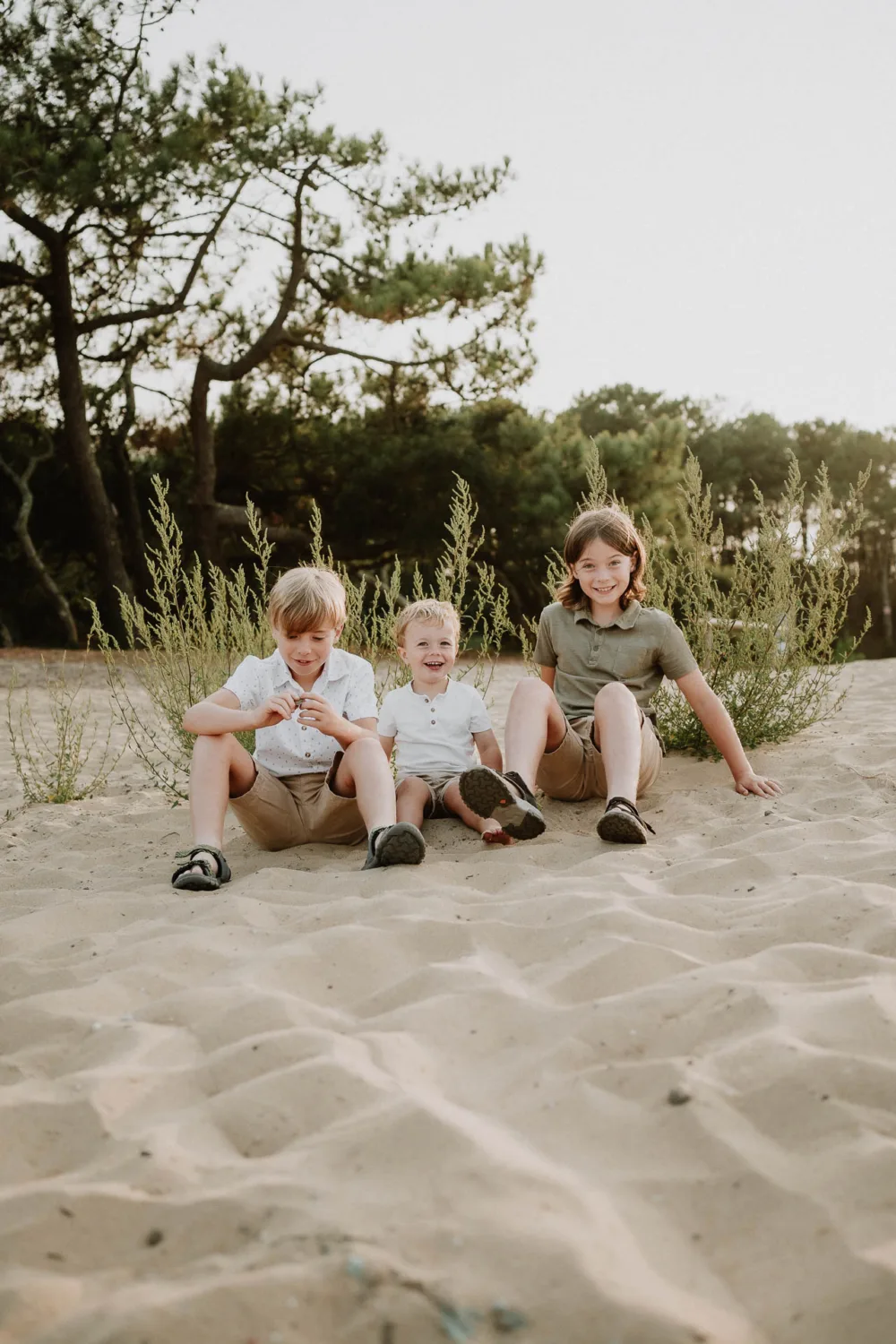 Sur cette image captivante optimisée pour le référencement en tant que photographe famille Landes, trois enfants - deux garçons et une fille - sont assis sur le sol sablonneux devant des buissons verts et des arbres, vêtus de tenues estivales décontractées tout en arborant de grands sourires, illustrant parfaitement la joie et la spontanéité typiques des séances photo de famille dans les magnifiques paysages naturels landais.