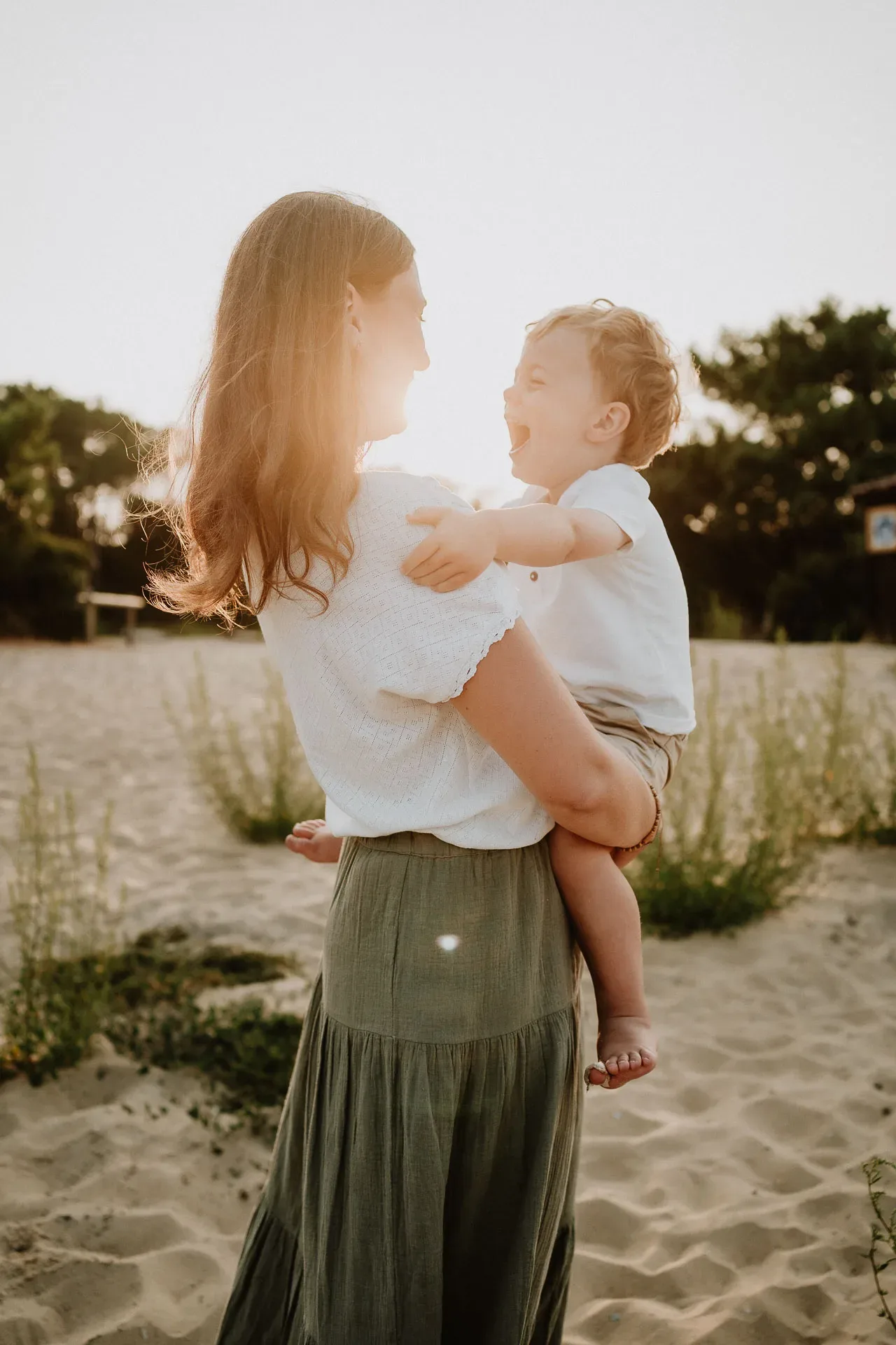 Une mère tient son enfant sur une plage de sable avec une verdure luxuriante en arrière-plan, les deux se regardant et échangeant un sourire chaleureux, immortalisés dans un moment tendre et authentique par un photographe de famille dans les Landes.