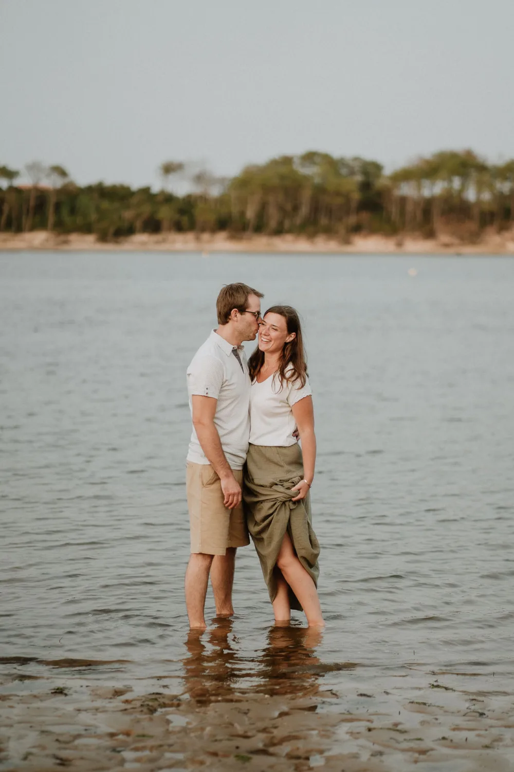 Un couple se tient dans l'eau peu profonde à la plage, l'homme embrasse tendrement la femme sur la joue tout en lui tenant la main, tous deux souriants et vêtus de vêtements décontractés d'été, avec des arbres en arrière-plan qui crée une scène idyllique typique des séances de photographe famille Landes.