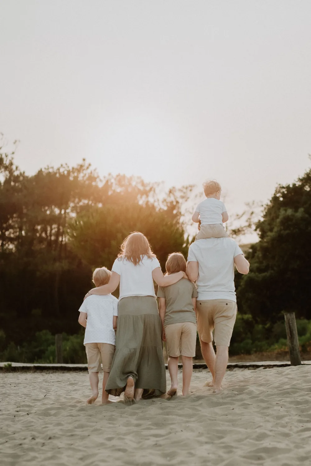 Un photographe de mariage ou de famille dans les Landes capturerait magnifiquement une famille de cinq personnes, composée de trois enfants, marchant main dans la main sur une plage sablonneuse au coucher du soleil dans les Landes, où le père porte le plus jeune enfant sur ses épaule tandis que des arbres et une végétation luxuriante font partie du décor enchanteur en arrière-plan.