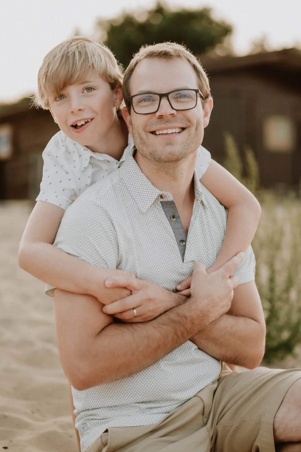 En tant que photographe de mariage et de famille dans les Landes, capturer des moments authentiques comme ce père portant des lunettes et une chemise à manches courtes, assister tout sourire tandis que son enfant aux cheveux blonds le serre tendrement dans ses bras en arrière-plan, est un pur bonheur ; cette scène en plein air avec sa verdure luxuriante et un bâtiment pittoresque à l'arrière-plan illustre à merveille la beauté naturelle des Landes et constitue une photo de famille idéale qui réchauffe les cœurs.