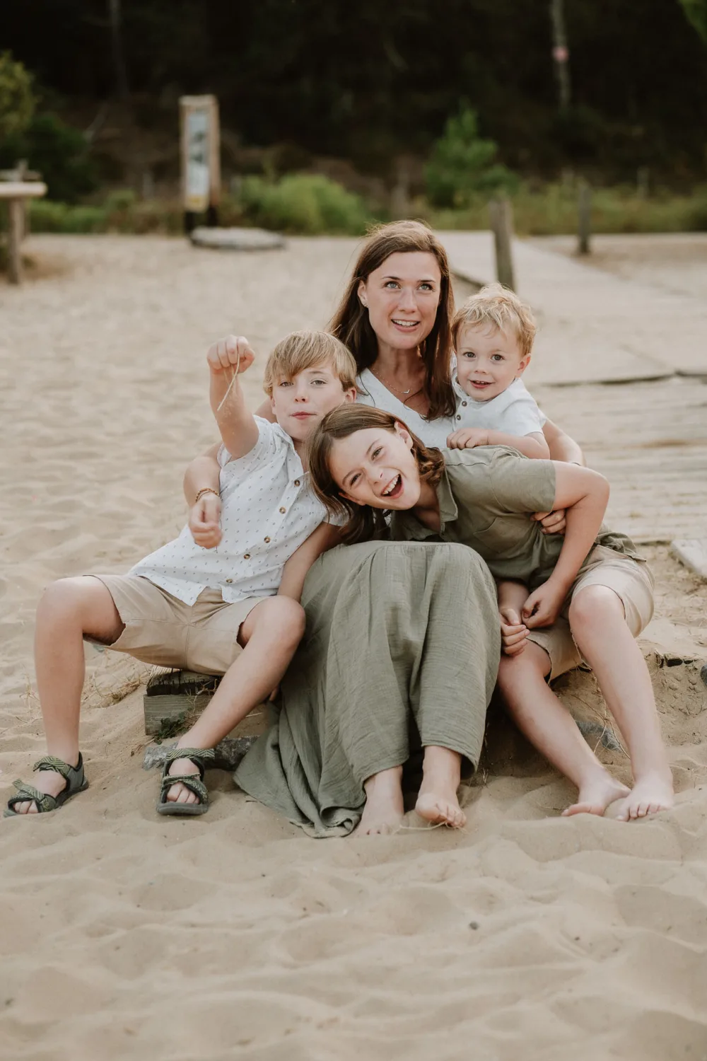 Une séance photo capturée par un photographe de famille dans les Landes montre une femme assise sur le sable, entourée de trois enfants – deux garçons et une fille – tous souriants et s'étreignant, créant ainsi un instant parfait rempli de bonheur familial sur une plage idyllique.