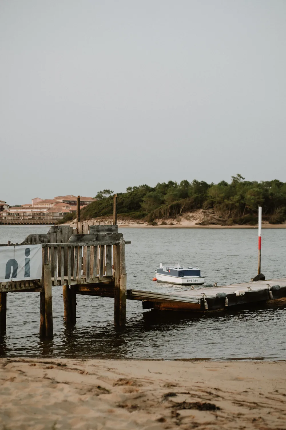 Sur une plage de sable dans les Landes, un petit bateau est amarré à un quai en bois surplombant une eau calme, idéal pour un photographe de mariage ou une famille cherchant à capturer des moments inoubliables avec en toile de fond une colline couverte d'arbres.