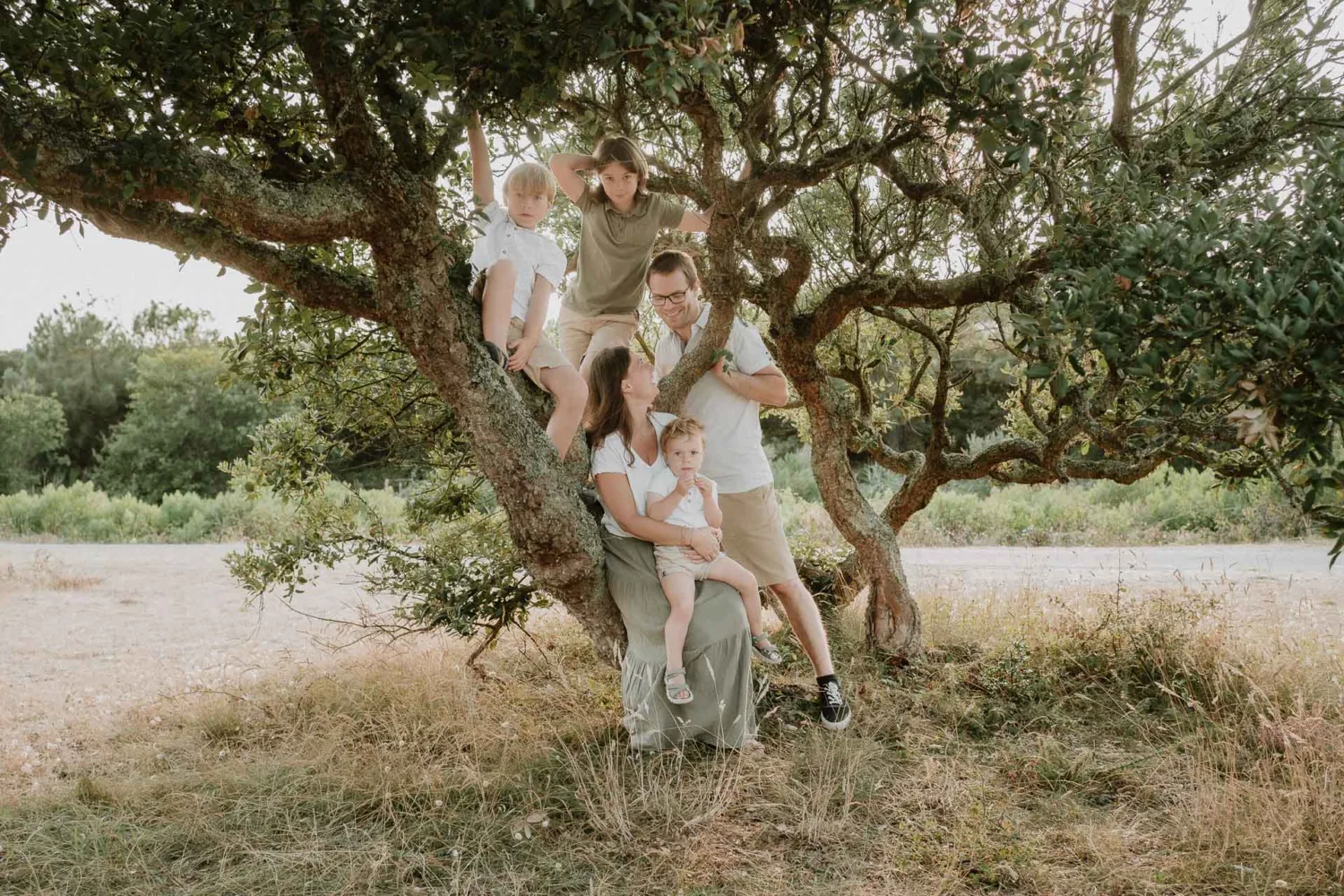 Une famille de cinq personnes, composée de deux adultes et trois enfants, réunie autour et sur un grand arbre dans un décor extérieur luxuriant avec une verdure apaisante en arrière-plan, magnifiquement immortalisée par Photographe Famille Landes pour créer des souvenirs inoubliables au cœur de la nature landaise.