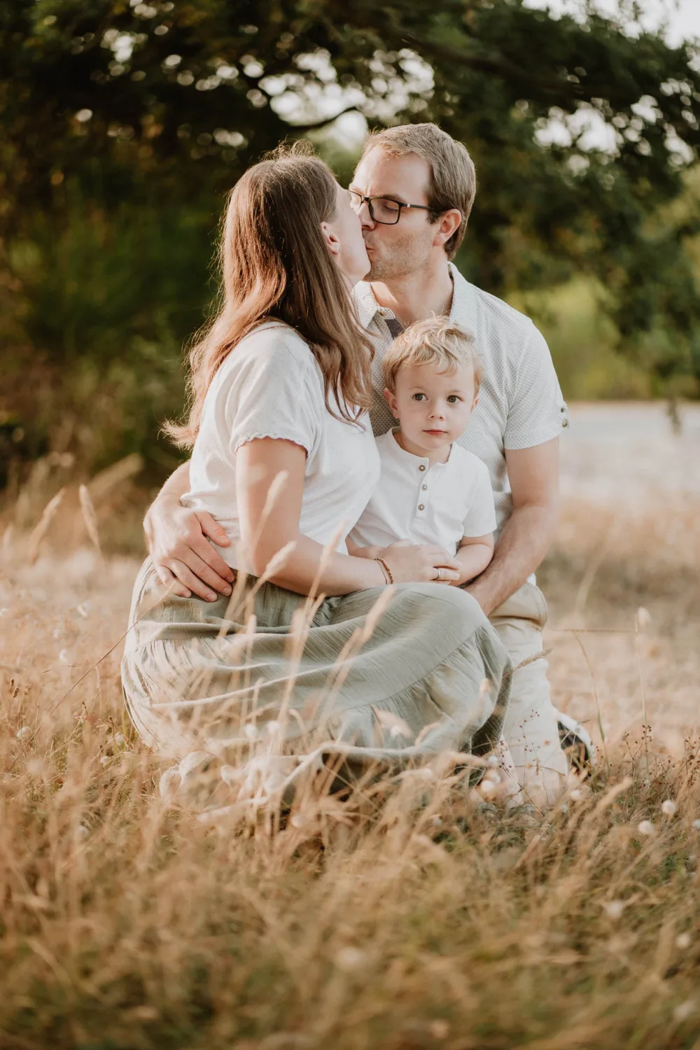 Cette image capturée par un photographe de famille dans les Landes montre une famille assise sur l'herbe en plein air, où un homme embrasse tendrement le devant d'une femme tandis que leur enfant regarde vers la caméra, tous les vêtements décontractés aux couleurs claires, avec de grands arbres se dressant gracieusement en arrière-plan.