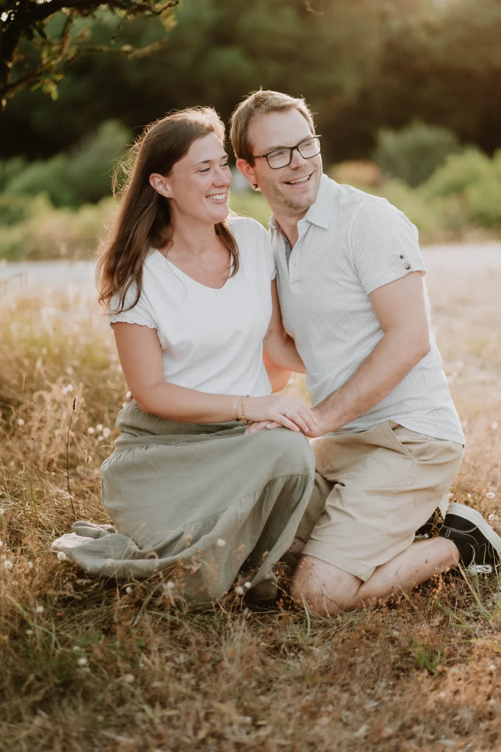 Un homme et une femme, habillés de manière décontractée, sont agenouillés sur un sol herbeux en plein air, souriant et regardant sur le côté, avec des arbres et du feuillage en arrière-plan évoquant un cadre paisible parfait pour une séance de photographe famille dans les Landes.