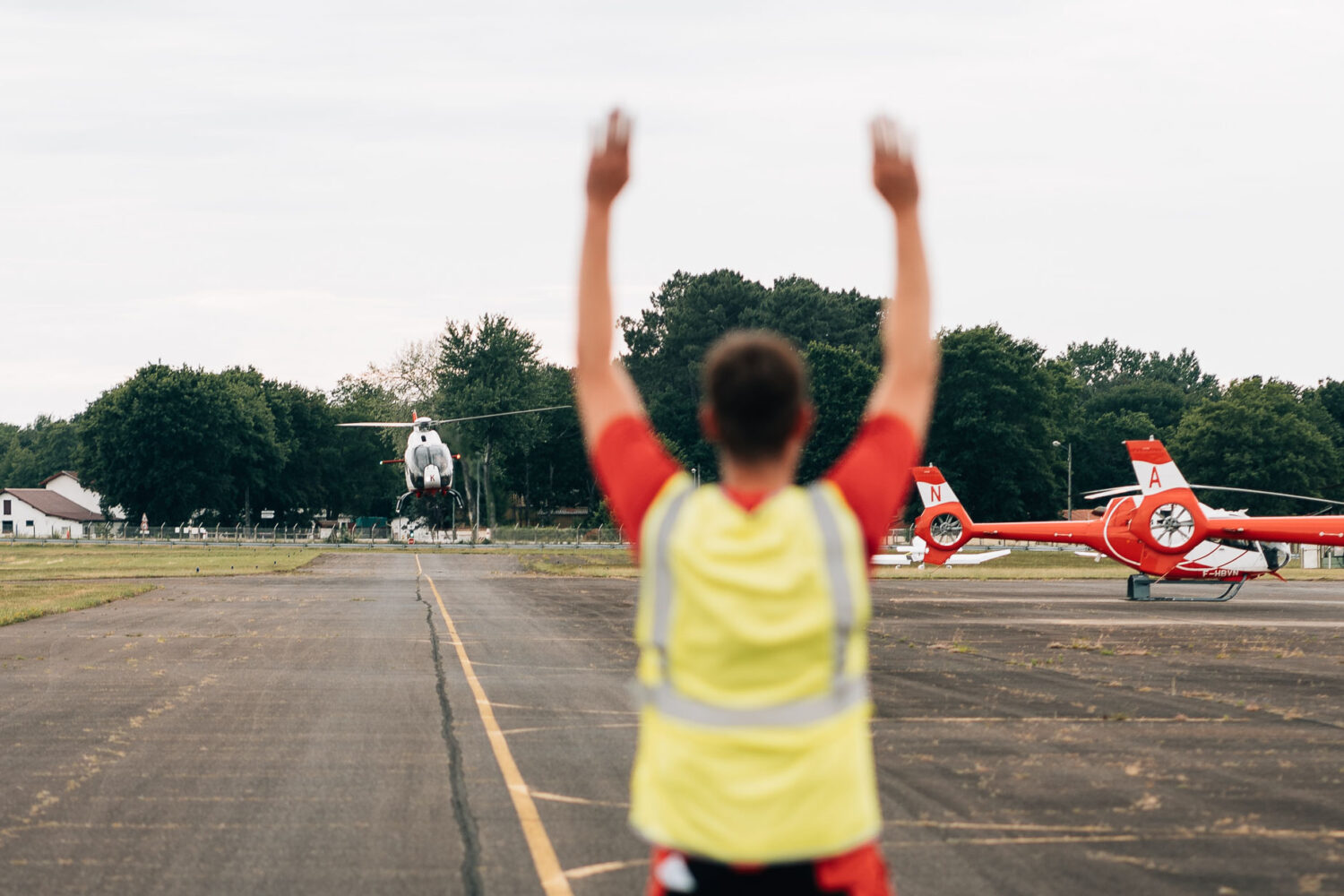 Cette image captivante, idéale pour un photographe de mariage ou de famille dans les Landes, montre une personne en gilet de sécurité qui guide un hélicoptère sur une piste d'atterrissage, avec pour toile de fond sereine deux autres hélicoptères stationnés, évoquant l'exaltant mélange d'adrénaline et de tranquillité que l'on ressent lors des vacances dans les Landes.