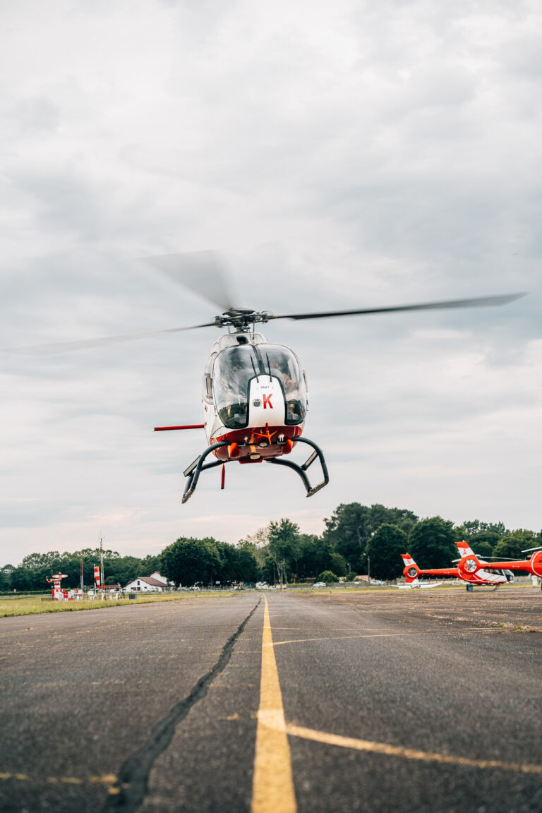 Un hélicoptère en vol stationnaire au-dessus d'une piste d'atterrissage, ses pales floues tranchant à travers le ciel nuageux, entouré d'arbres et d'autres hélicoptères stationnés, incarne l'esprit aventurier des vacances dans les Landes tout en capturant l'excitation du voyage même par une journée terne, offrant un cadre parfait pour immortaliser des moments inoubliables de photographie de mariage ou en famille dans le cadre naturel enchanteur et dynamique des Landes.