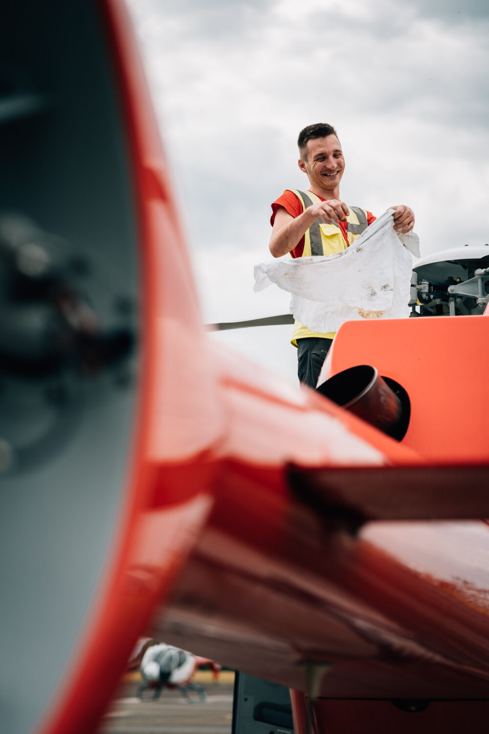 Sur cette image optimisée pour le référencement, un professionnel de la photographie de mariage ou de famille dans les Landes capture une scène unique où une personne vêtue d'un gilet de sécurité nettoie délicatement à l'aide d'un chiffon un avion orange brillant, le tout se détache élégamment sur un ciel nuageux évoquant la beauté sereine et pittoresque des vacances dans les Landes, lieu pris pour son charme naturel et ses décors enchanteurs distinctifs pour immortaliser des moments inoubliables en famille ou lors d'une cérémonie nuptiale.