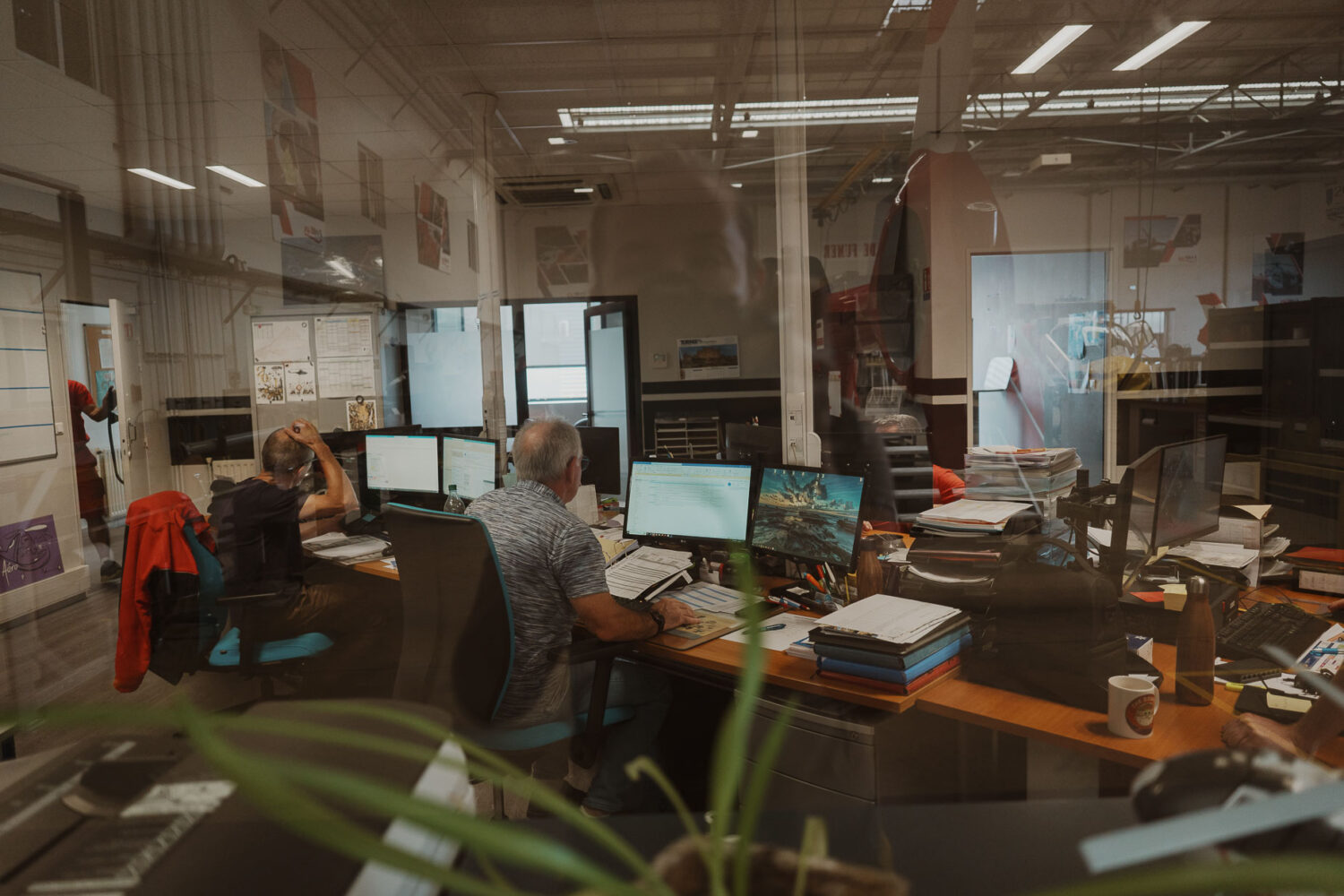 Derrière la paroi vitrée, un bureau bourdonne d'activité alors que plusieurs personnes travaillent sur des ordinateurs, leurs bureaux encombrés de papiers et de multiples écrans, offrant un contraste frappant avec les rêves d'une séance photo de mariage ou famille dans les Landes où la sérénité et les ciels ouverts remplacent les écrans numériques et les courriels sans fin.