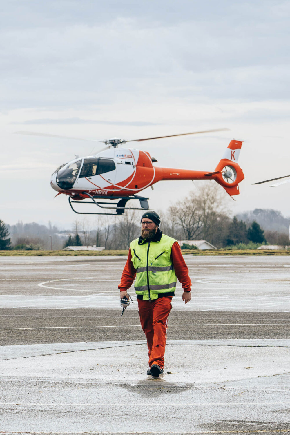 Sur le tarmac d'un aéroport pittoresque des Landes, un homme vêtu d'un gilet haute visibilité et de pantalons rouges déambule en arrière-plan d'un hélicoptère rouge et blanc, capturant l'esprit aventureux des vacances dans les Landes tout en offrant une opportunité photographique unique pour les portraits de mariage ou de famille, idéalement intégrée dans le paysage naturel et vibrant de cette région emblématique du sud-ouest de la France.