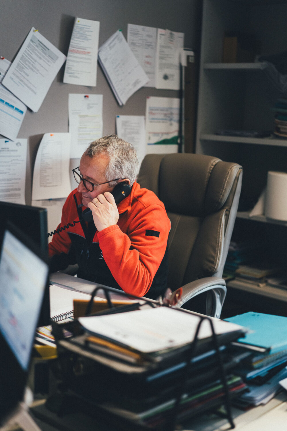 Sur cette image, un homme d'un certain âge portant des lunettes et une veste rouge et noire est capturé en pleine conversation téléphonique, assis à un bureau encombré de papiers et de fournitures de bureau, probablement en train d'évoquer ses futures vacances familiales. ou un mariage dans les Landes, une destination prisée pour ses paysages pittoresques qui inspirent de nombreux photographes spécialisés en photographie de mariage et de famille.