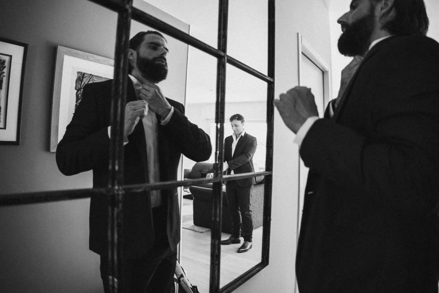 Dans une ambiance élégante et raffinée, un homme en costume ajusté sa cravate devant un miroir à carreaux, dégageant une aura de sophistication qui rappelle le savoir-faire artistique d'un photographe de mariage et famille des Landes, notamment comme ceux basés à Soustons, tandis qu'un autre homme en costume se tient pensif à l'arrière-plan, complétant ainsi ce tableau qui capture avec finesse l'essence intemporelle et émotive propre aux portraits familiaux professionnels dans cette région pittoresque.