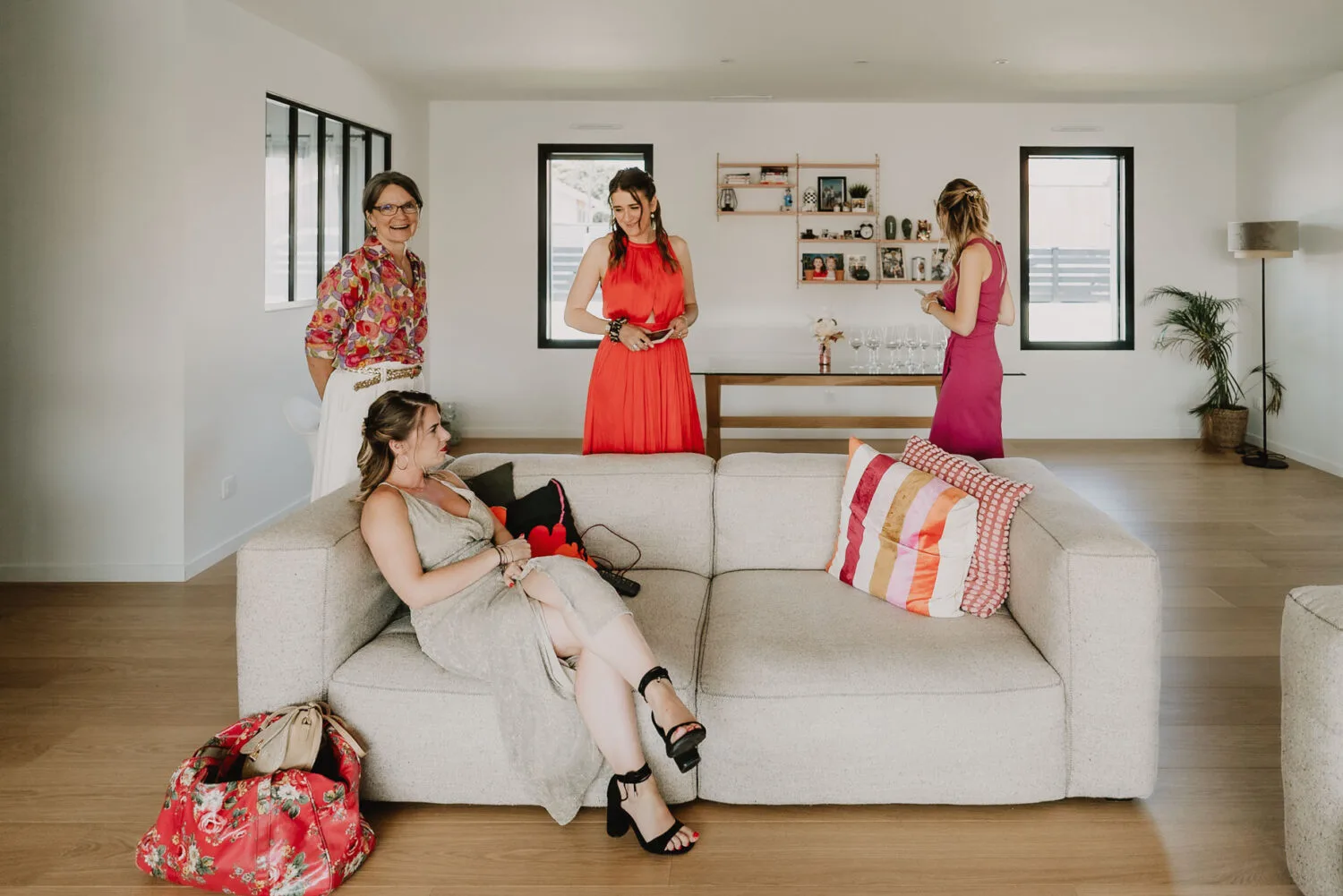 Dans un salon lumineux à Soustons, une scène intime capturée par un photographe de mariage et de famille des Landes révèle quatre femmes réunies pour un portrait familial serein ; l'une est assise sur le canapé pendant que les trois autres se tiennent debout à proximité, entourées d'une atmosphère douce créée par la lumière tamisée filtrante à travers deux petites fenêtres qui éclairent subtilement une étagère ornée de décorations murales.