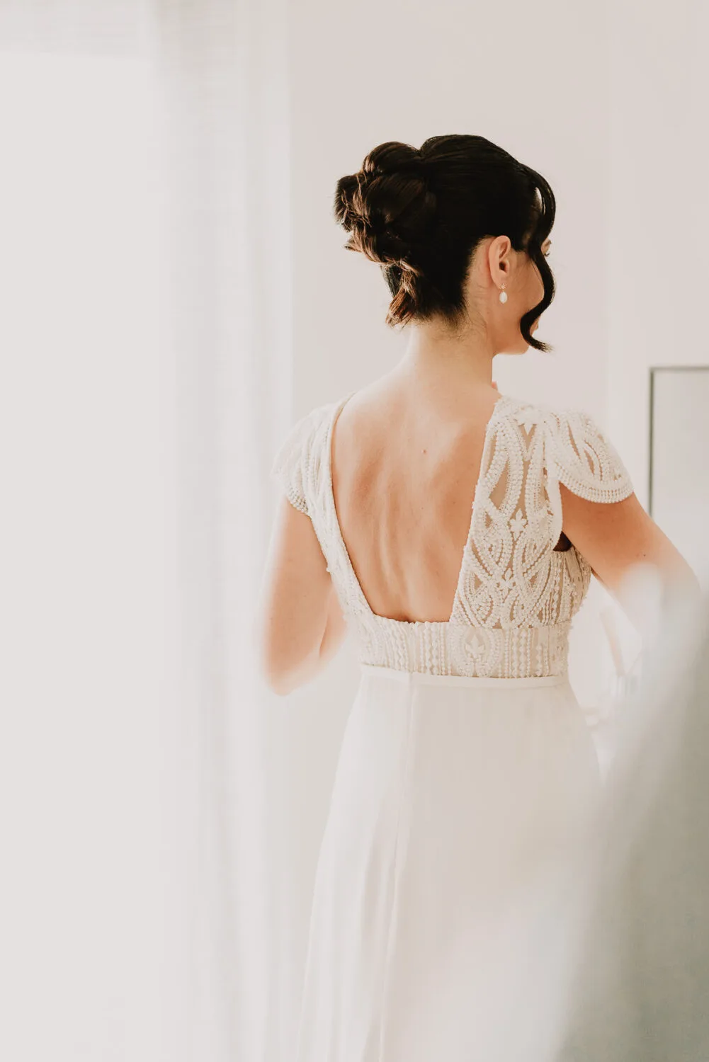 Dans cette image évoquant le style distinctif d'un photographe de mariage et de famille dans les Landes, tout particulièrement inspiré par l'esthétique des séances photo à Soustons, une femme est vêtue d'une élégante robe blanche ornée de délicats motifs en dentelle se tient dos à une grande et lumineuse fenêtre, capturant un moment de sérénité et d'intimité qui symbolise la beauté intemporelle des souvenirs familiaux.