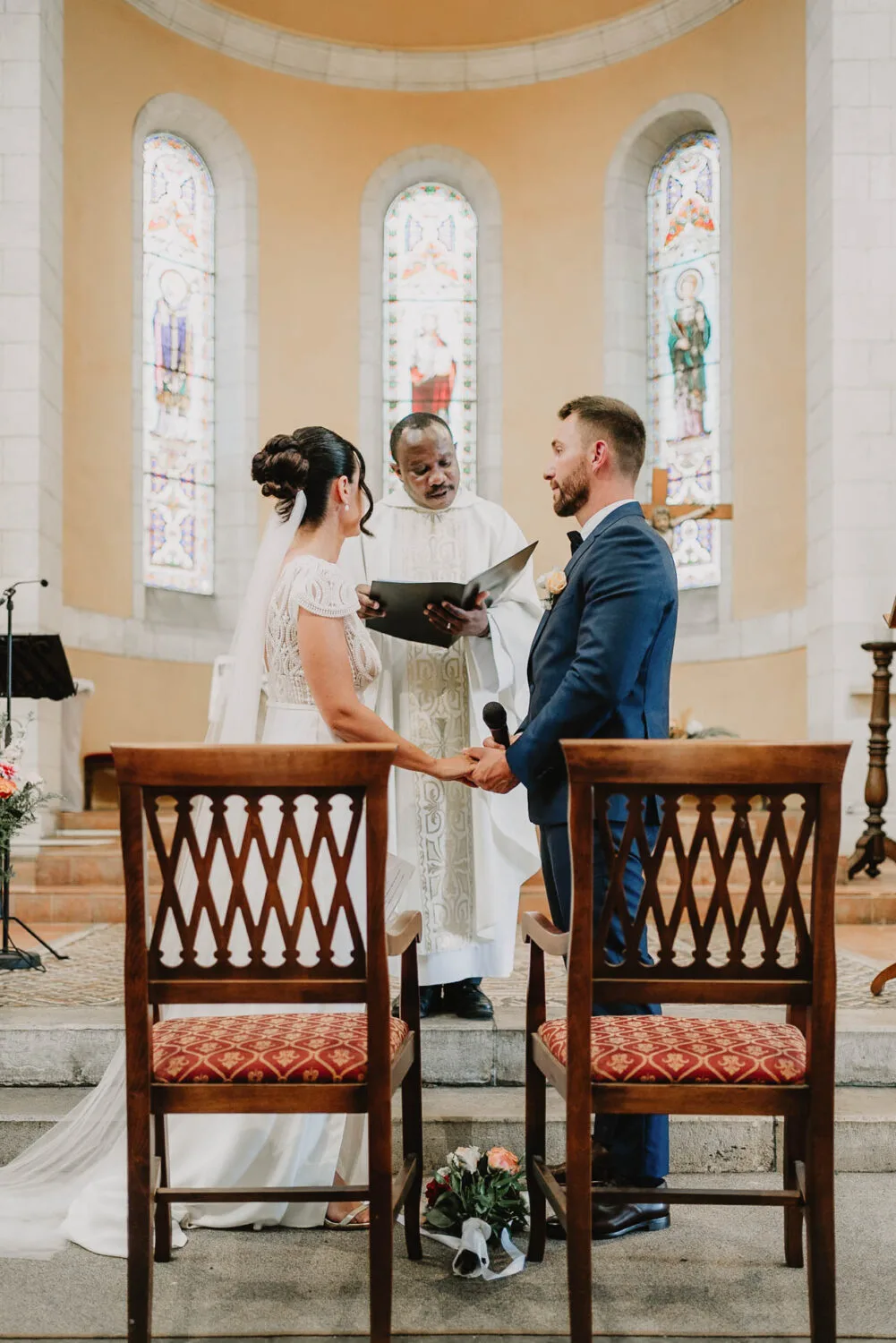 Lors d'une cérémonie de mariage émouvante dans une charmante église des Landes, un couple de mariés se tient la main, tandis qu'un prêtre officie leurs vœux ; deux chaises ornées de coussins rouges ajoutent une touche chaleureuse à l'avant-plan, capturant parfaitement l'esprit intime de cet événement idéal pour tout photographe de mariage ou photographe de famille Soustons cherchant à immortaliser ces instants précieux.