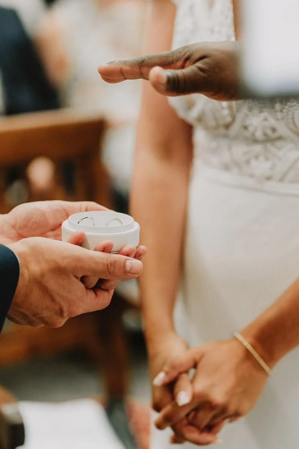 Cette image saisie de près l'instant intime d'une cérémonie de mariage, où une bague est présentée dans un écrin ouvert tandis que deux mains se serrent amoureusement en arrière-plan, un moment précieux que tout *photographe mariage et famille des Landes* à Soustons rêverait d'immortaliser grâce à son expertise en capturant ces connexions authentiques et émouvantes.