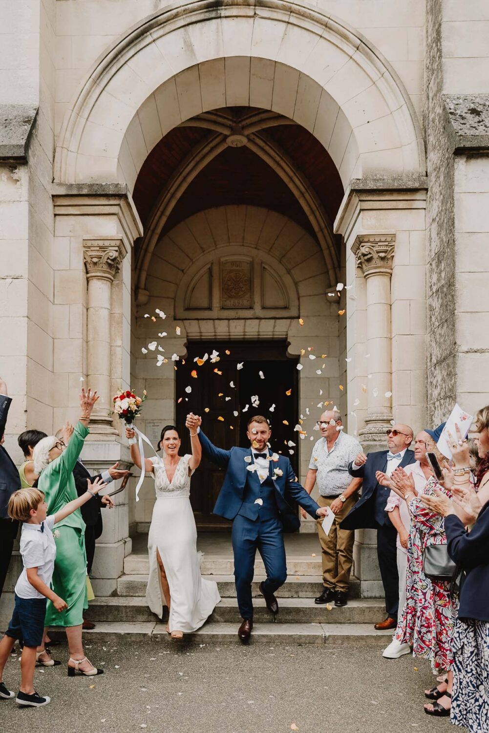 Un couple de jeunes mariés rayonne de bonheur en sortant main dans la main d'un bâtiment en pierre à Soustons, tandis qu'un photographe de mariage talentueux capture cet instant précieux entouré d'invités lançant du confetti, immortalisant ainsi un souvenir inoubliable dans le Cadre enchanteur des Landes.