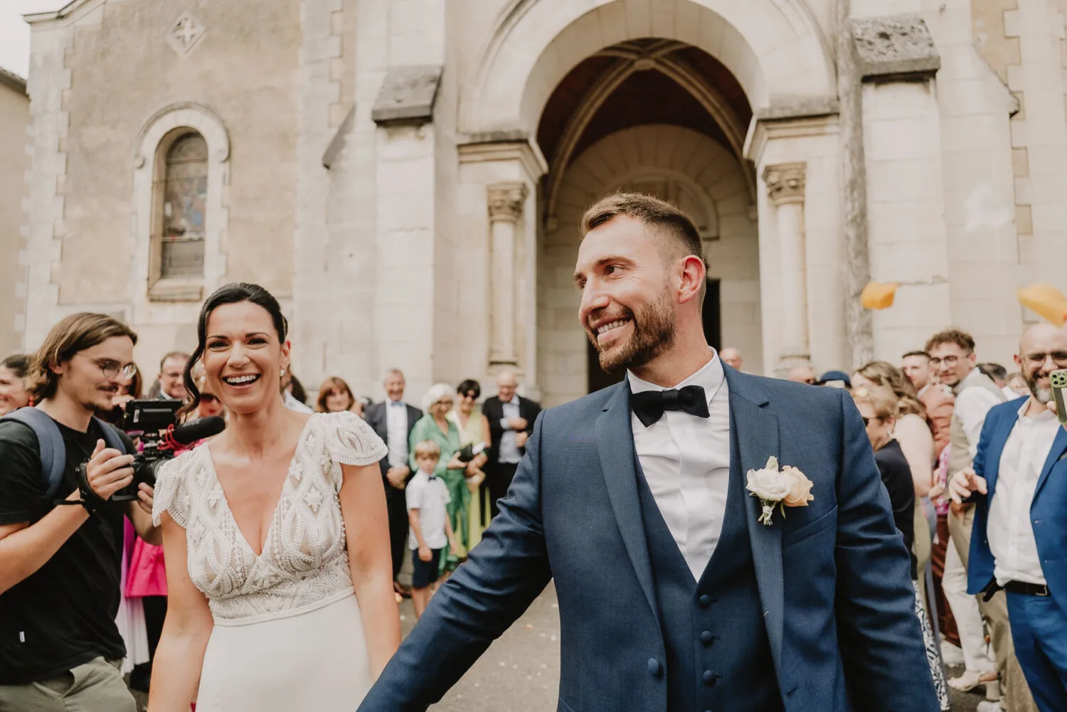 Devant un bâtiment de pierre dans les pittoresques Landes, un couple de mariés rayonne de bonheur en se tenant la main, tandis qu'une foule d'invités enthousiastes se presse derrière eux en immortalisant l'événement sur leurs appareils photo ; c'est l'instant idéal pour un *photographe mariage Soustons* ou même un *photographe famille Soustons* de saisir toute la joie et l'amour qui emplissent l'air.