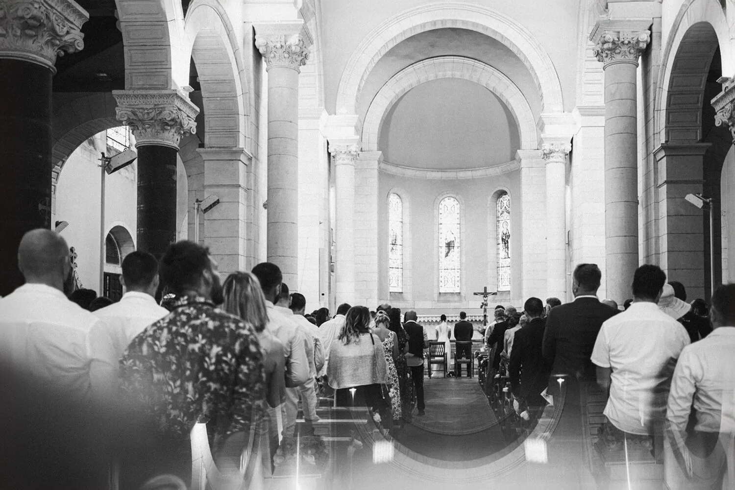 Cette photo en noir et blanc immortalise une cérémonie de mariage dans une église, où l'assemblée est assise face à l'autel, et reflète parfaitement le style distinctif du photographe de famille à Soustons avec son architecture voûtée et ses fenêtres en vitraux magnifiques à l'arrière-plan, capturant ainsi le charme intemporel des mariages dans les Landes.