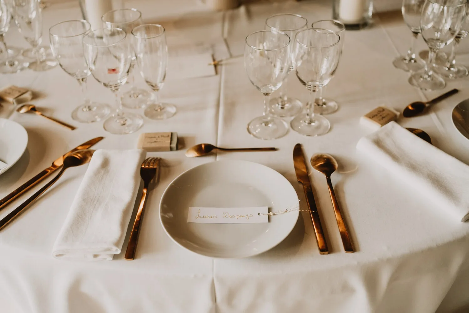 Cette scène raffinée, parfaite pour un photographe de mariage et de famille dans les Landes, met en vedette un décor de table élégant avec des assiettes blanches immaculées, des couverts dorés étincelants et des serviettes méticuleusement pliées sur une nappe blanche, entourées de nombreux verres à vin et cartes de placement soigneusement disposées rappelant l'art photographique d'une ambiance familiale à Soustons.