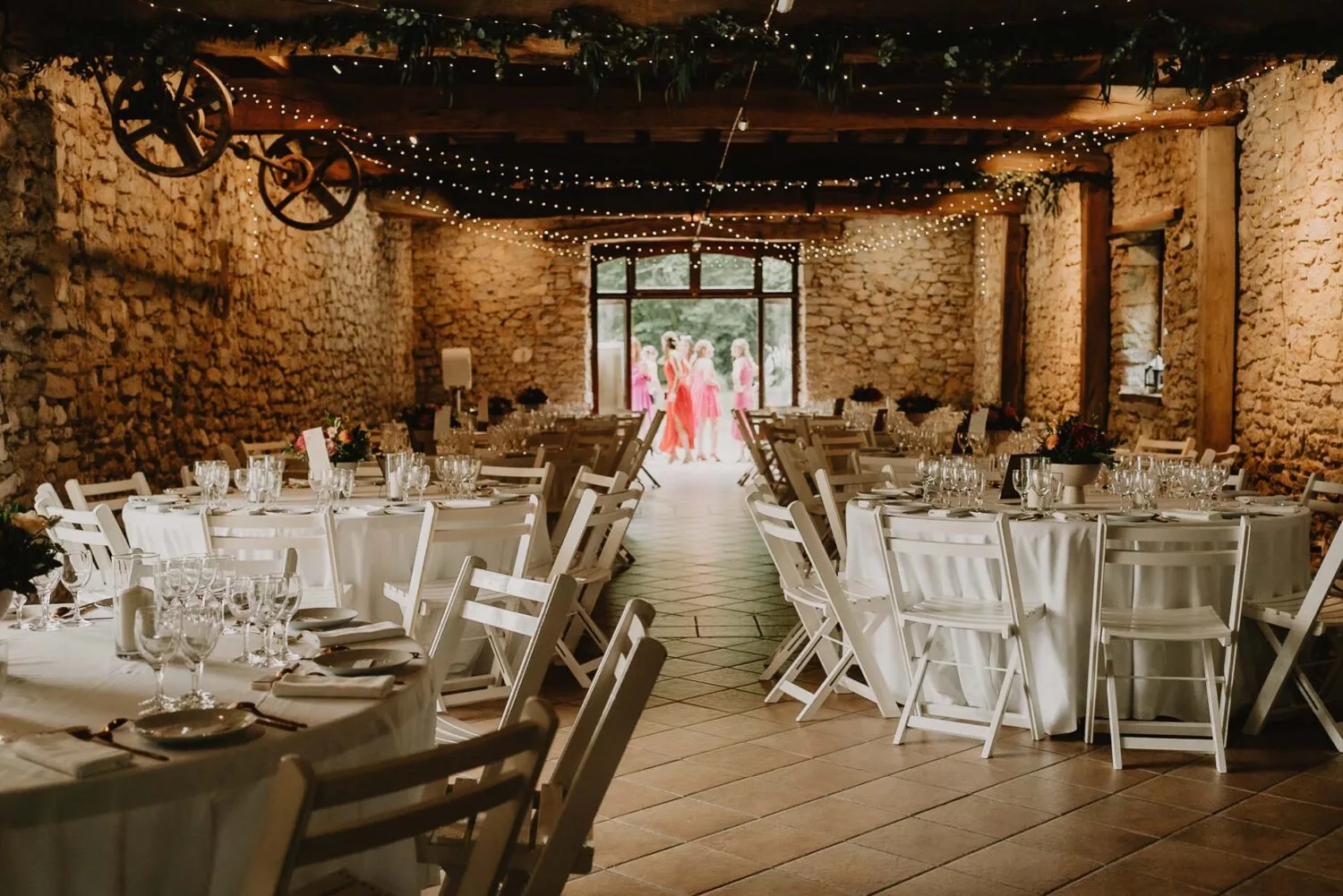 Dans cette image captivante optimisée pour le référencement, on découvre une salle de réception de mariage au charme rustique dans les Landes, ornée de murs en pierre décorées et éclairée d'une lumière tamisée créant une ambiance intimiste ; les tables rondes sont élégamment dressées pour le dîner et, à l'arrière-plan, un groupe de personnes se tient près de l'entrée, rappelant une scène authentique généralement immortalisée par un photographe de famille à Soustons.