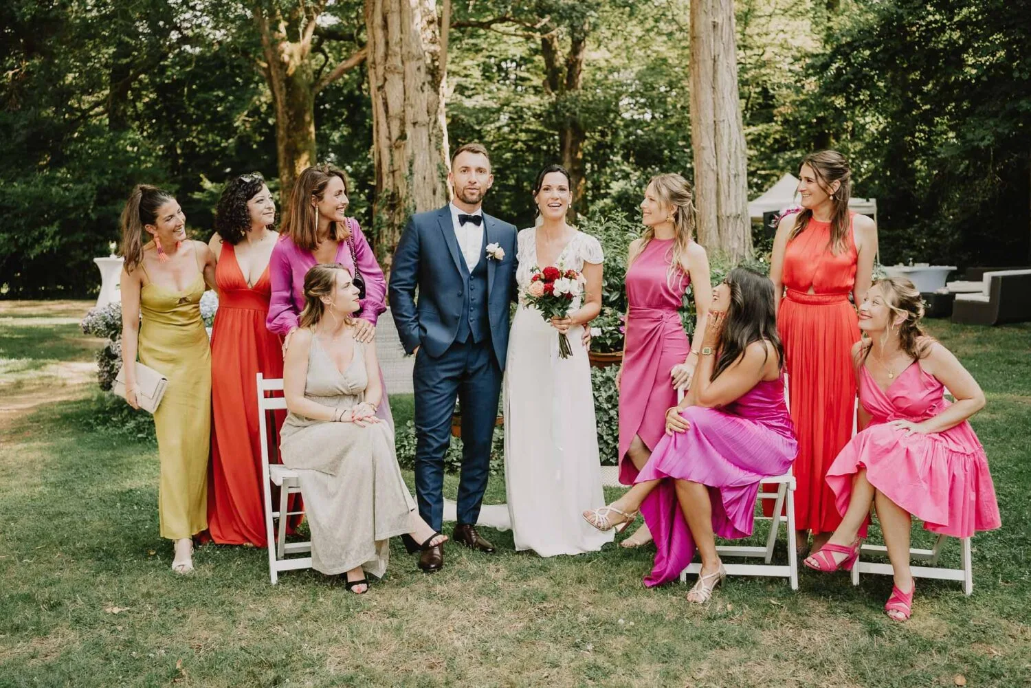 Dans un cadre enchanteur idéal pour les prestations d'un photographe de mariage ou de famille dans les Landes, une mariée et un marié porteront avec un groupe de femmes vêtues de robes éclatantes, certaines assises et d'autres debout, sur fond luxuriant d'une forêt verdoyante à Soustons, offrant ainsi une scène parfaite pour immortaliser des souvenirs intemporels.