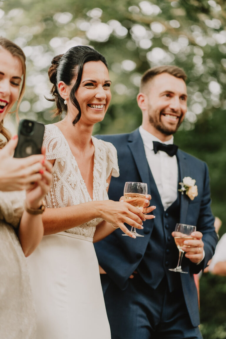 Sur cette image captivante optimisée pour le référencement, un couple de mariés en tenue élégante sourit chaleureusement tout en tenant des verres de vin, entouré de joyeux invités lors d'un mariage inoubliable dans les Landes; ce moment empreint de bonheur et d'émotion est immortalisé avec l'expertise d'une talentueuse famille photographe basée à Soustons, garantissant des souvenirs impérissables qui resteront gravés dans les mémoires.