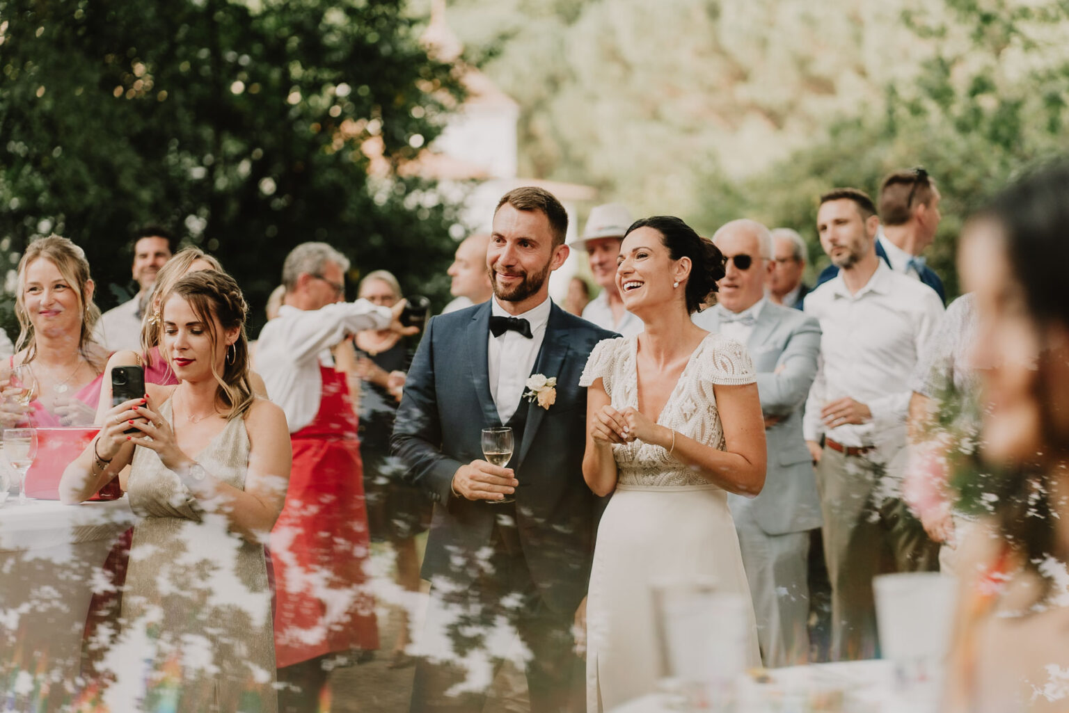 Au cœur d'une célébration en plein air à Soustons, une mariée et un marié se tiennent côte à côte avec un radieux, entourés de leurs invités lors de cette réception festive familiale dans les Landes, tandis que certains convives savourent des boissons et qu 'une femme capture cet instant mémorable, le tout sous l'œil attentif d'un photographe spécialisé en mariages et événements familiaux chargé d'immortaliser ce moment de bonheur.