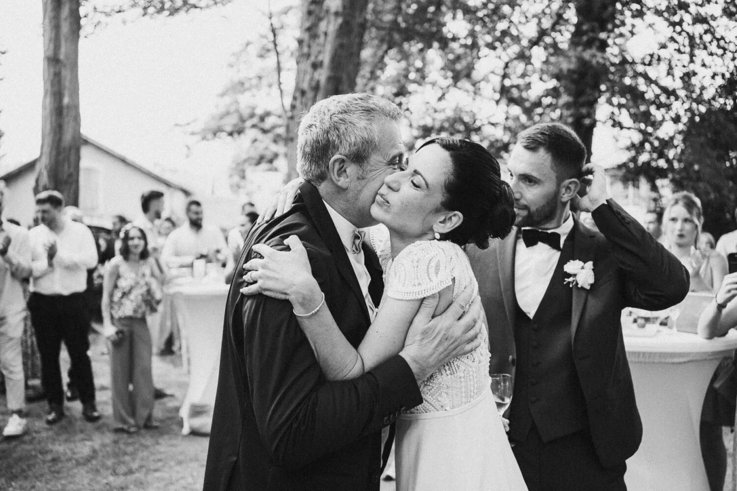 Lors de la réception en plein air d'un mariage mémorable immortalisé par le talentueux photographe famille Soustons, une mariée étreint chaleureusement un homme plus âgé sous le regard bienveillant du marié à ses côtés, tandis qu'en arrière-plan, les invités et les Les tables décorées témoignent de l'atmosphère conviviale et joyeuse typique des célébrations dans les Landes.