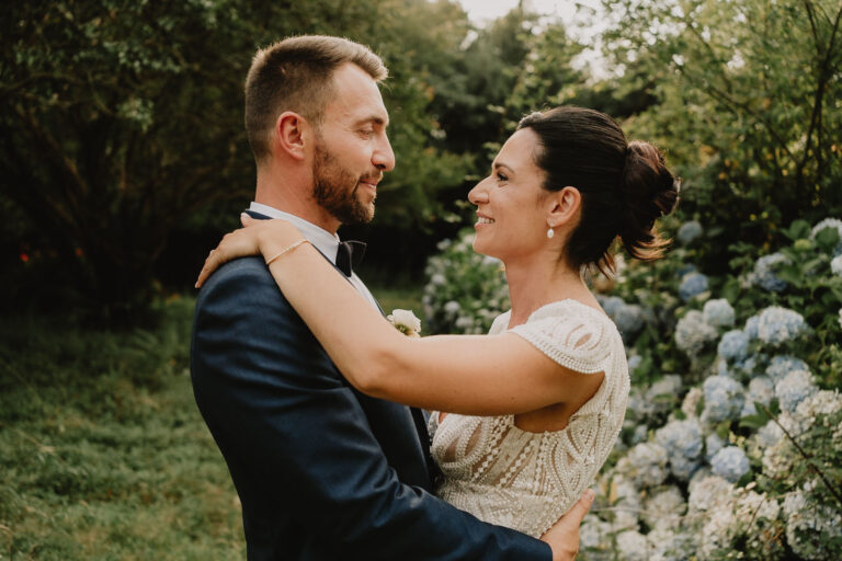 Dans cette scène enchanteuse digne d'une collection de photographe mariage et famille dans les Landes, un couple s'enlace tendrement en plein air, entouré d'une verdure luxuriante et de fleurs d'hortensia éclatantes, vêtus élégamment pour capturer la magie intemporelle de l'amour à Soustons, où leurs sourires radieux apportent une complicité profonde et authentique.