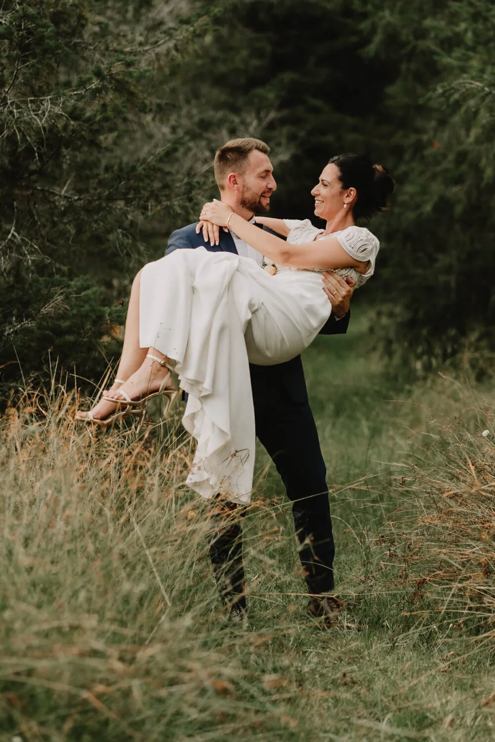 Un marié vêtu d'un costume sombre porte sa mariée souriante habillée d'une robe blanche à travers un champ verdoyant entouré d'arbres majestueux, illustrant parfaitement la joie authentique et intemporelle que tout photographe de mariage ou de famille dans les Landes, notamment le photographe famille Soustons, s'efforce de saisir lors de ces moments uniques.