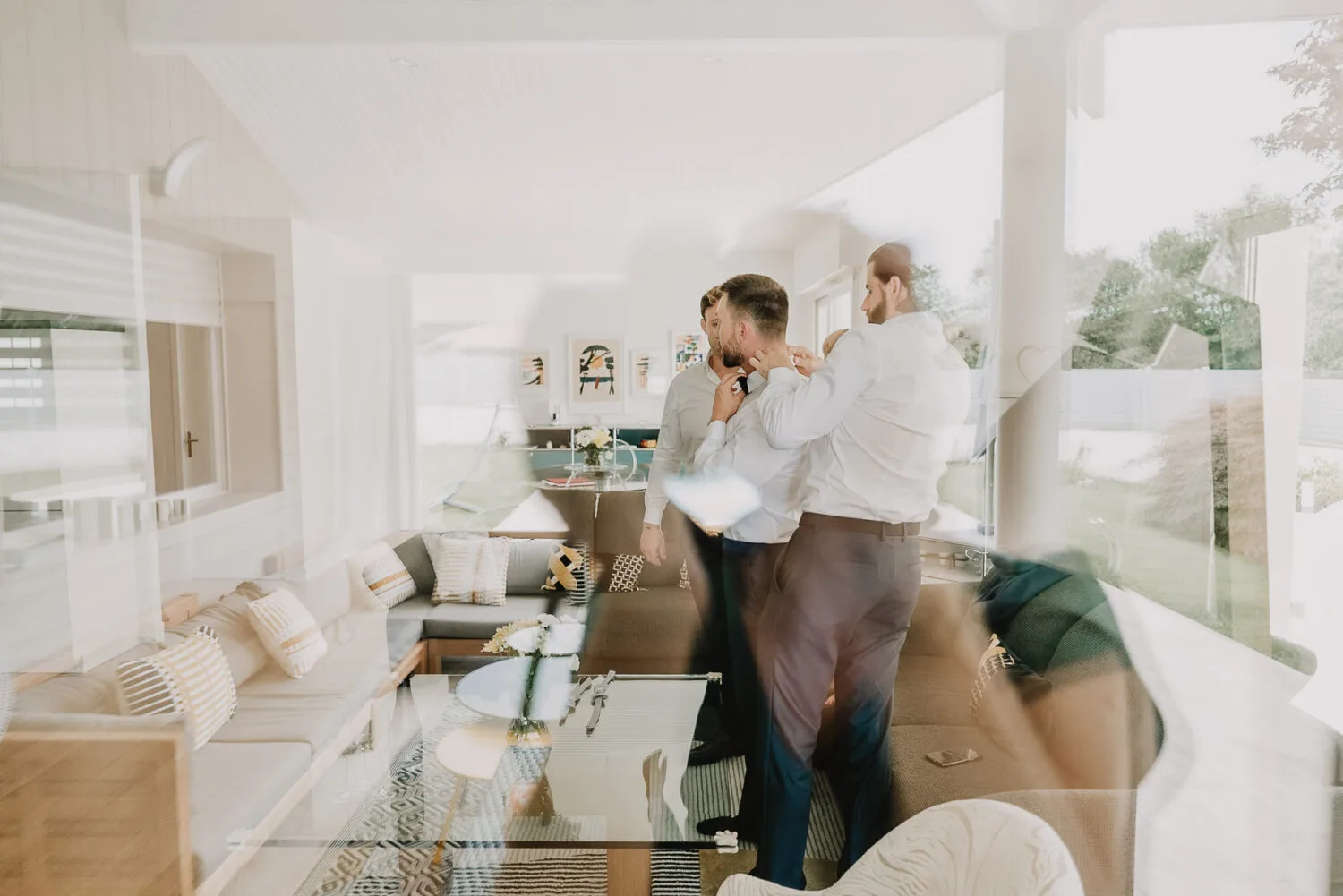 Dans un salon lumineux et moderne aux vastes fenêtres illuminant un canapé beige, deux hommes en chemises ajustent leurs cravates, capturant l'élégance et la complicité idéale pour une séance photo réalisée par un photographe de mariage ou de famille à Soustons, dans les Landes.