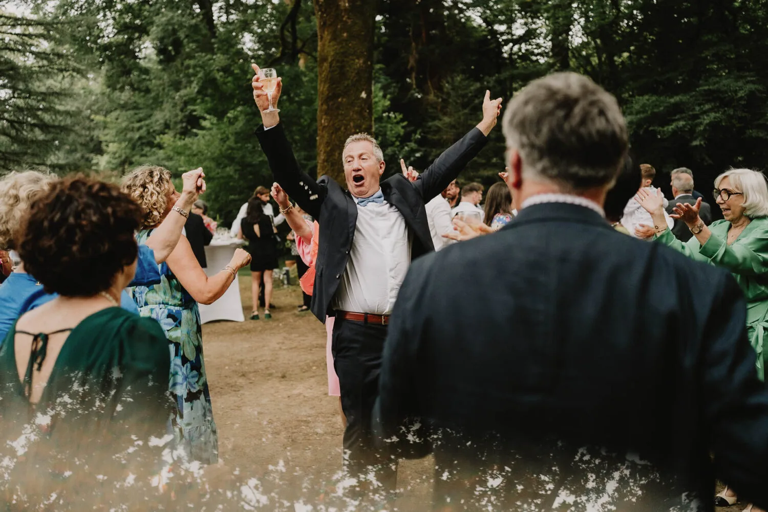 Au cœur d'une ambiance festive typique d'un photographe mariage Soustons, un homme enthousiaste lève les bras et son verre dans une célébration joviale, entouré de nombreux convives qui se tiennent autour de lui lors d'une réunion en plein air dans la beauté boisée naturelle des Landes, capturant l'essence même de ces moments précieux que seul un expert en photographie de famille peut immortaliser.