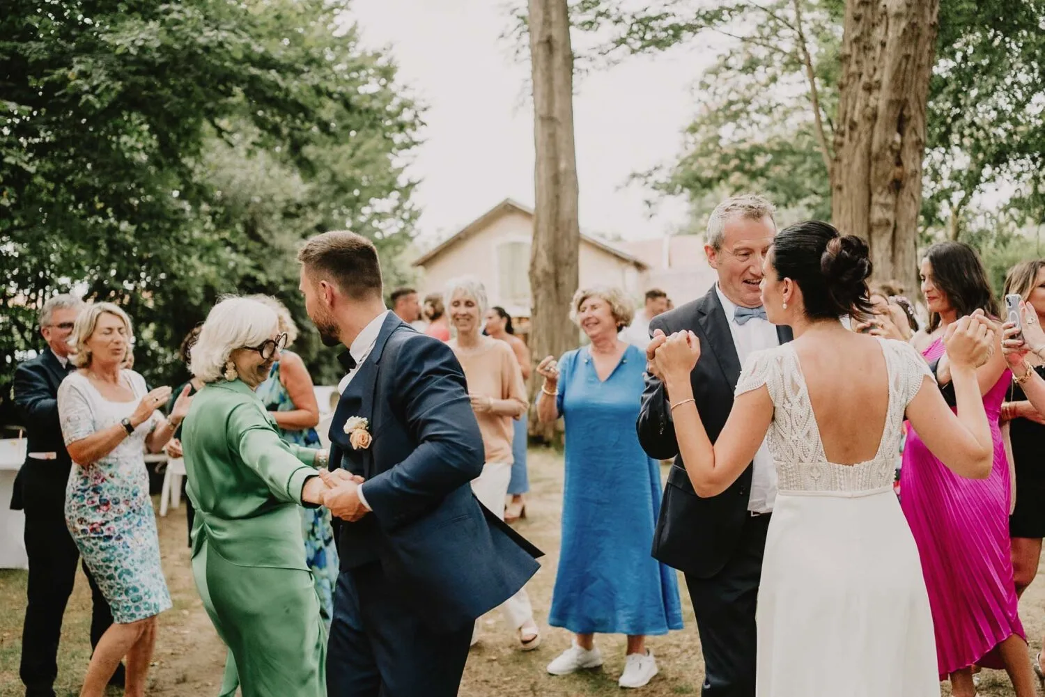 Lors d'une réception de mariage en plein air dans les magnifiques Landes, où la nature se mêle harmonieusement à la célébration, deux couples en tenue de soirée élégante dansent avec grâces entourées d'invités vêtus de couleurs vives, sous le regard attentif des arbres majestueux et d'un bâtiment pittoresque en arrière-plan, offrant un cadre idéal pour capturer des moments inoubliables grâce au talent d'un photographe de famille spécialisé à Soustons.