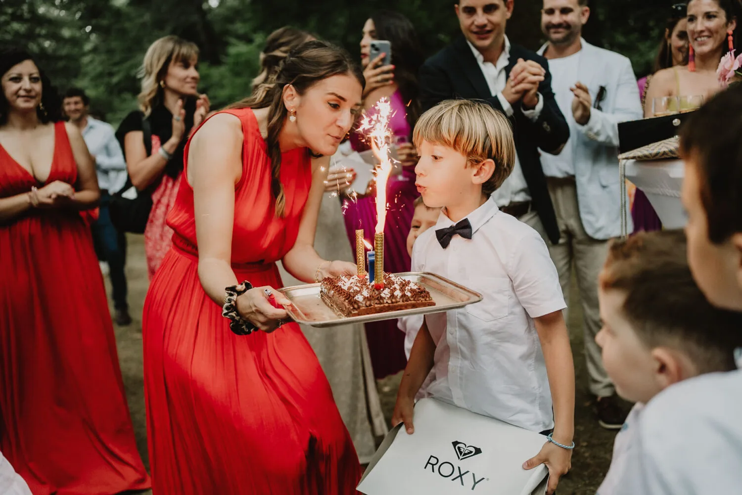 Dans cette image captivante capturée par un photographe de famille à Soustons, une femme en robe rouge, incarnant l'élégance typique des moments festifs dans les Landes, tient un gâteau orné de cierges magiques scintillants prêt à être soufflés par un jeune garçon portant un nœud. papillon, tandis que leurs proches en habitudes de fête forment une toile de fond joyeuse et attentive pour immortaliser cet instant familial précieux et chaleureux.