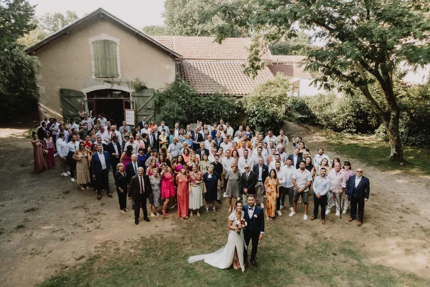 Cette magnifique photo de groupe de mariage prise en extérieur devant un bâtiment rustique capture un couple élégamment vêtu au centre, entouré d'invités vêtus de tenues colorées, et reflète à merveille l'amour et la joie d'une célébration authentique dans une atmosphère champêtre pleine de charme; réalisée par un photographe mariage et famille à Soustons dans les Landes, cette image illustre parfaitement l'art de capturer des moments précieux en pleine nature landaise.