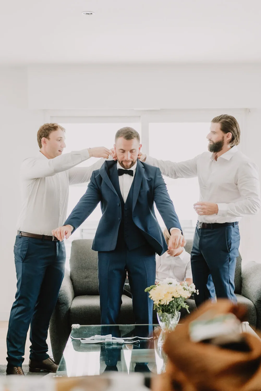 Dans une salle de séjour lumineuse, trois hommes s'activent autour du marié pour ajuster son costume et son nœud papillon impeccablement, tandis qu'un photographe de famille à Soustons immortalise ce moment précieux en plein cœur des Landes, devant une table en verre ornée d'un élégant vase fleuri qui apporte une touche de sophistication à cette scène pré-nuptiale chaleureuse.