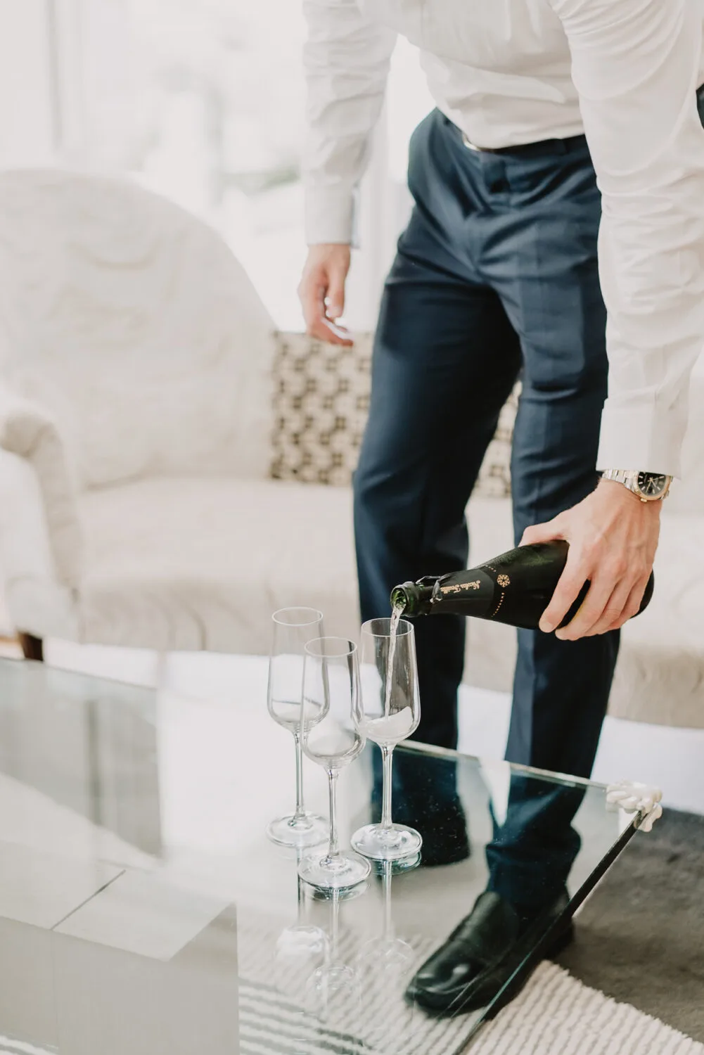 Dans un salon élégant, évoquant le cadre enchanteur d'une séance de photographe de mariage ou de famille dans les Landes à Soustons, une personne vêtue avec raffinement vers gracieusement du champagne dans trois flûtes étincelantes posées sur la table en verre, créant une atmosphère conviviale et sophistiquée idéale pour des souvenirs intemporels.