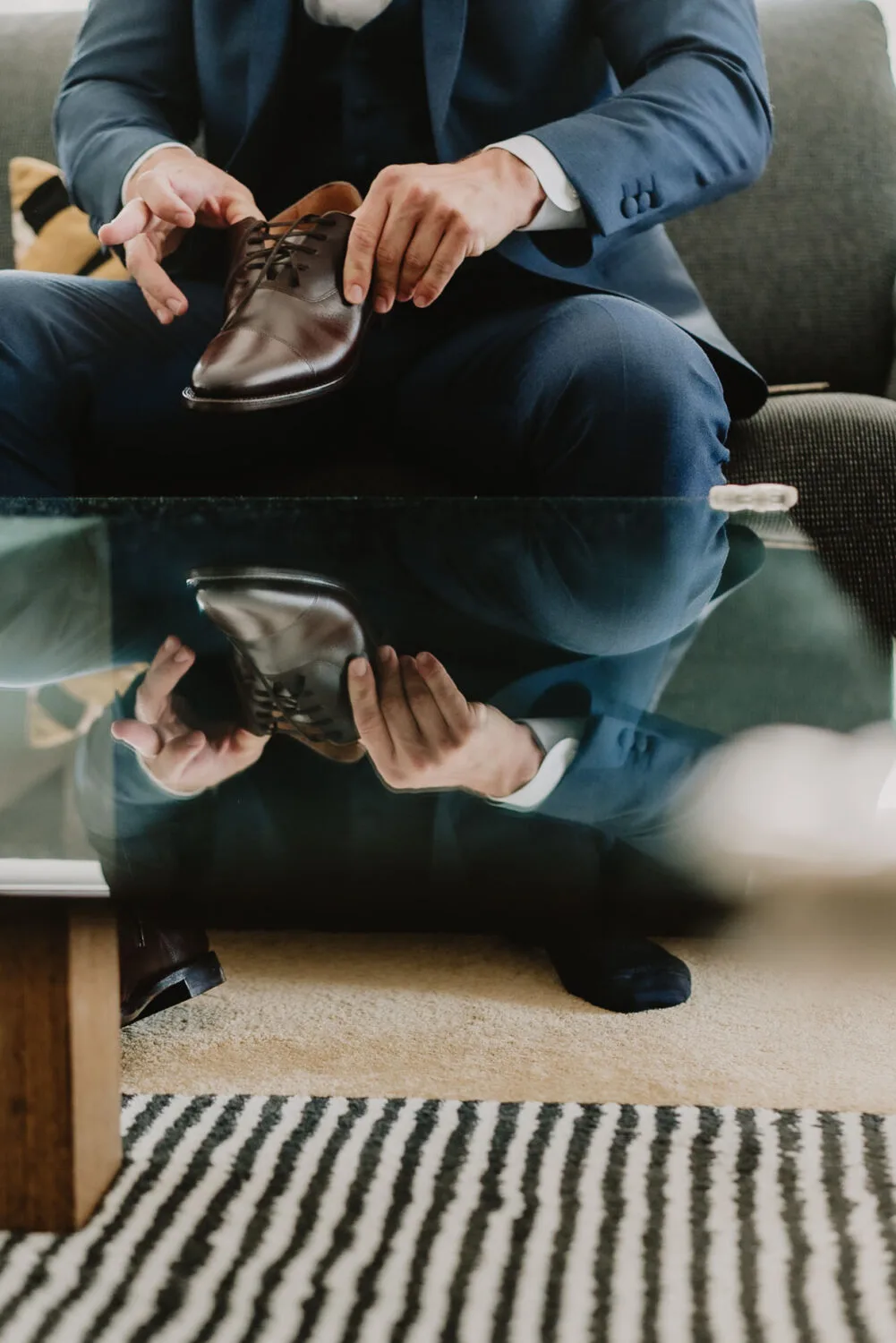 Dans une ambiance élégante et soignée typique d'un photographe de mariage ou famille des Landes, un individu en costume est assis sur un canapé tout en tenant une chaussure habillée marron au-dessus d'une table en verre, le tout posé sur un tapis. rayé qui évoque le charme rustique et chaleureux propre aux séances photo en famille à Soustons.