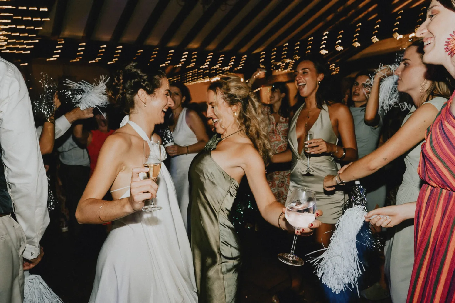 Un groupe animé de personnes danse et fête en intérieur, tenant des boissons et des pompons sous des guirlandes lumineuses scintillantes—une ambiance festive qui pourrait être parfaitement immortalisée par un photographe expert en mariage ou famille dans les Landes, capturant chaque moment précieux avec créativité et sensibilité.