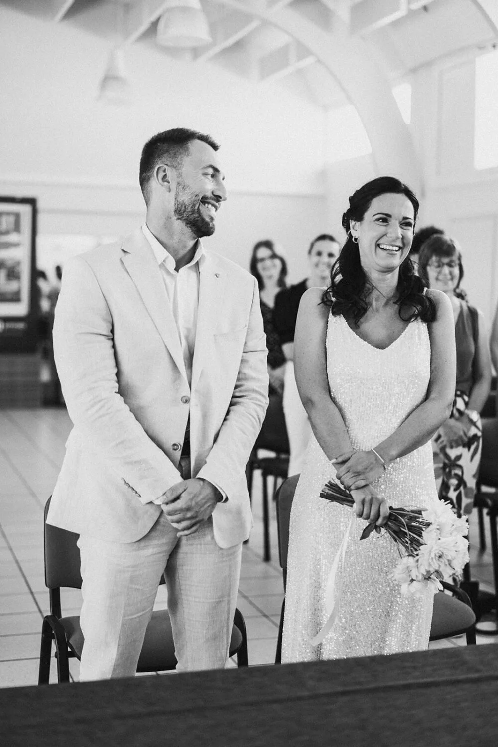 Au cœur d'une cérémonie élégante immortalisée par un photographe de mariage talentueux des Landes, un couple rayonnant en tenue formelle se tient debout à l'intérieur, la femme tenant un bouquet tandis que l'atmosphère chaleureuse est rehaussée par plusieurs invités assis en arrière. -plan, offrant ainsi une parfaite illustration de la magie des souvenirs capturés à Soustons.