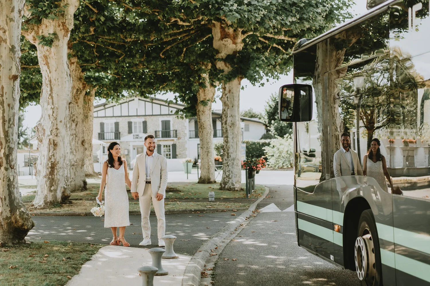 Sur une pittoresque rue bordée d'arbres à Soustons, au cœur des Landes, un couple vêtu de vêtements clairs se tient main dans la main à côté d'un grand bus, immortalisant un instant de quiétude idéal pour un photographe de mariage ou famille recherchant des scènes authentiques et paisibles dans cet écrin naturel parfait.