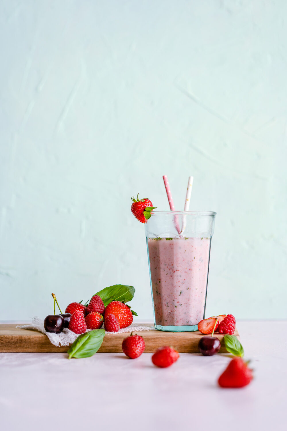 Cette image captivante, mise en scène par un photographe professionnel basé dans les Landes, présente de manière artistique un verre de smoothie rose orné de deux pailles, entouré délicatement de fraises et de cerises posées sur une planche à découper, le tout se détachant magnifiquement sur un fond vert clair, illustrant parfaitement le talent et l'originalité d'un photographe spécialisé en mariages et séances photo familiales dans cette région pittoresque.