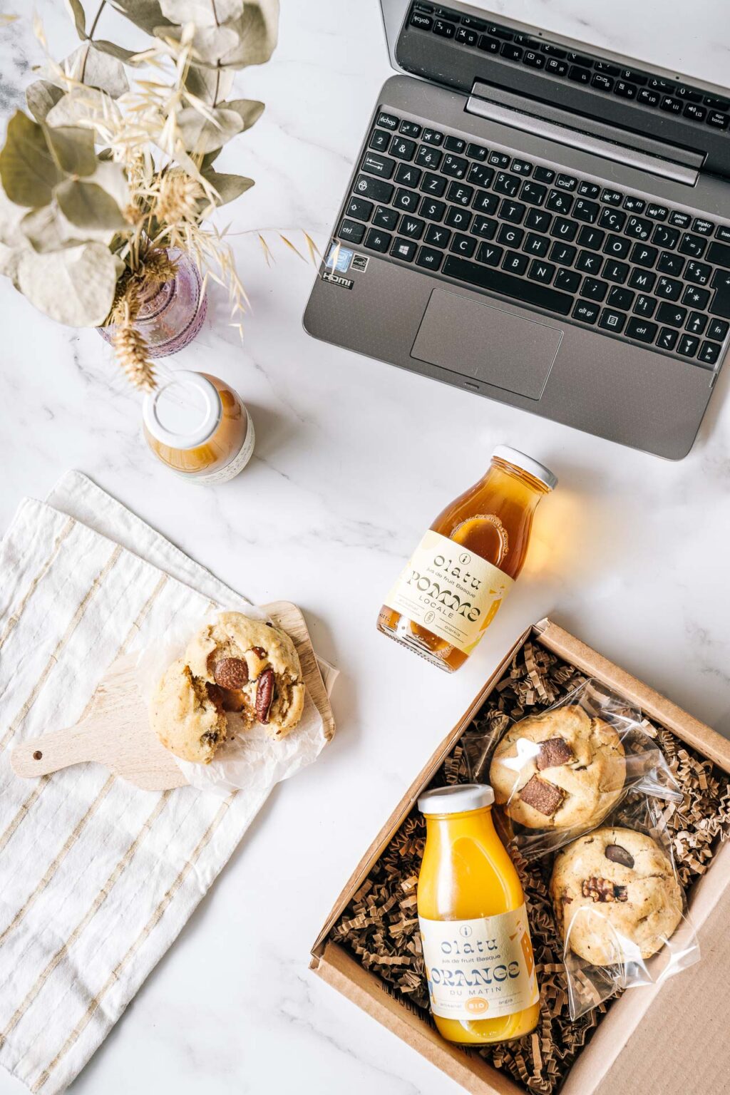 Cette image, réalisée par un photographe professionnel spécialisé dans les mariages et séances photo de famille dans les Landes, met en scène un élégant ordinateur portable posé sur une surface en marbre, entouré d'éléments chaleureux et accueillants comme des bouteilles de jus d'orange rafraîchissant, une boîte remplie de délicieux cookies prêts à être dégustés, une planche en bois ornée d'un cookie gourmand pour une touche rustique, le tout agrémenté par un linge rayé qui apporte mouvement et texture à la composition visuelle harmonieuse rehaussée par des fleurs séchées artistiquement placées dans un vase.